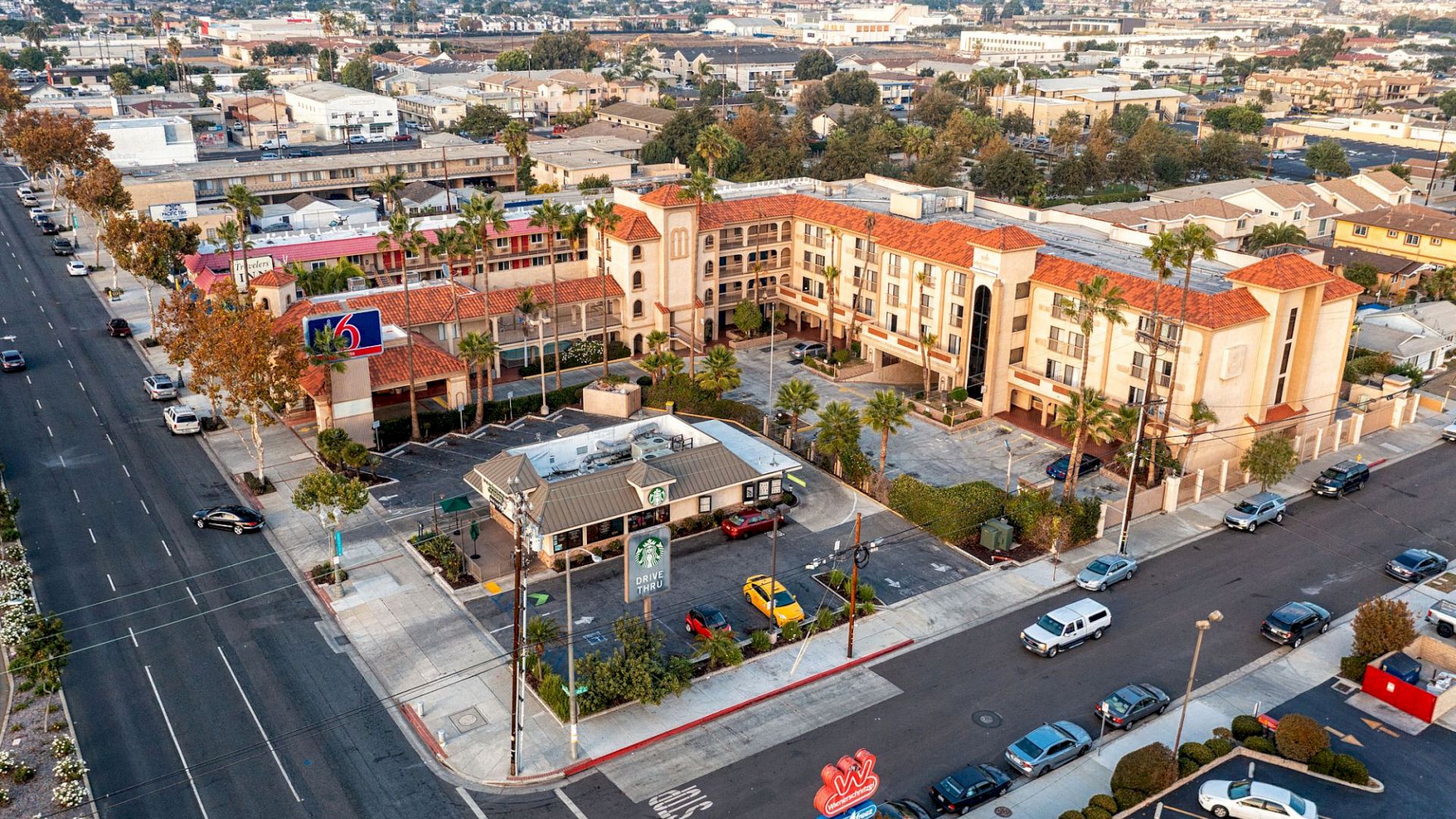 An aerial view of an urban area showing buildings, roads, and cars. A multi-story hotel and a small eatery with a parking area are visible in the foreground.