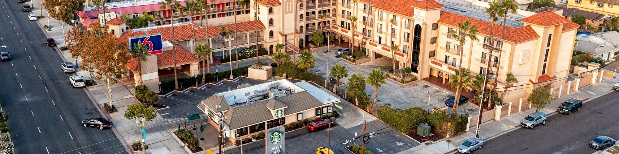 An aerial view of an urban area showing buildings, roads, and cars. A multi-story hotel and a small eatery with a parking area are visible in the foreground.