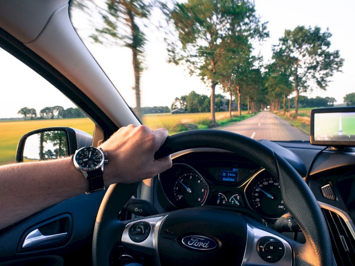 The image shows a person driving a Ford car down a tree-lined road, with a visible watch on their wrist and a GPS device mounted on the dashboard.