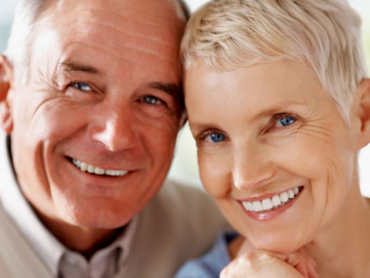 An elderly couple is smiling at the camera. They look happy and content. Both of them are wearing light-colored clothing. They are close together.