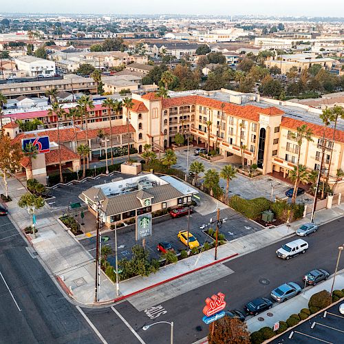 An aerial view of a street with buildings, including a hotel, a few restaurants, and parking lots, surrounded by a residential area in the background.