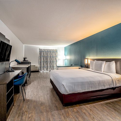 A modern hotel room with a large bed, a flat-screen TV, a desk, a chair, nightstands, and stylish lighting.