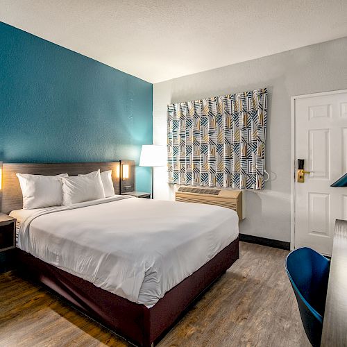 A modern hotel room with a double bed, blue accent wall, desk, chair, lamp, and patterned curtains, featuring a mix of wood and white decor.