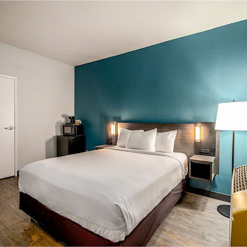 A modern hotel room with a double bed, teal accent wall, side tables, a standing lamp, small appliances, and a patterned curtain.