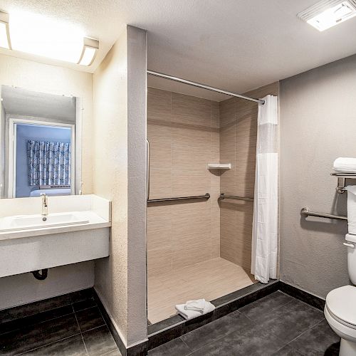 A clean bathroom with a sink, large mirror, shower area with curtain, and a toilet with towel racks and folded towels.