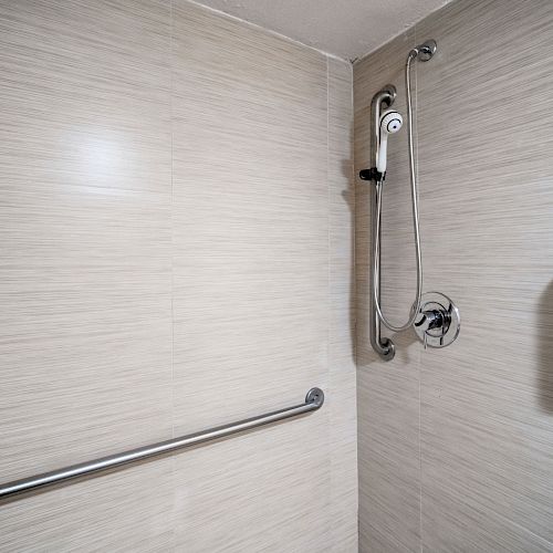 The image shows a shower area with beige tile walls, a handheld shower head, and two stainless steel grab bars mounted on the wall.