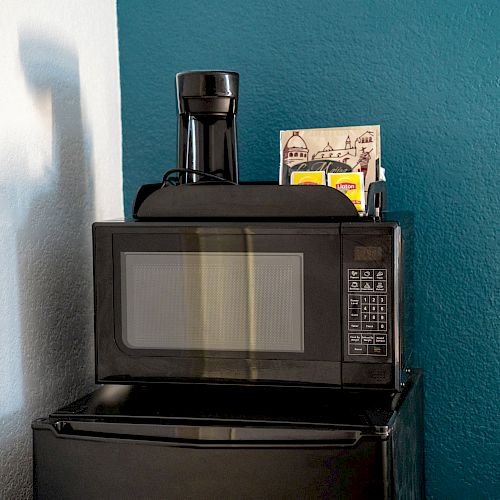 A black microwave oven is placed on top of a small fridge, with a black coffee maker and some papers or magazines on top of the microwave.