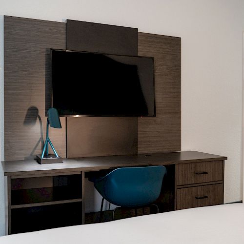 A room with a wall-mounted TV, a desk, a blue chair, a lamp, and a closed door can be seen.