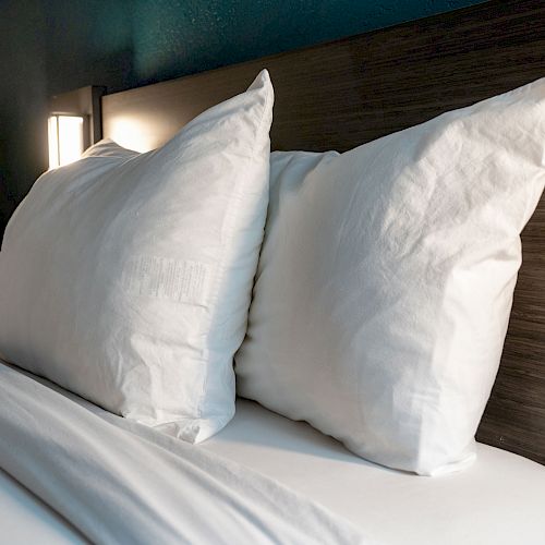 A neatly made bed with white pillows and sheets, a wooden headboard, and a nightstand with a coffee maker and cups in the background.