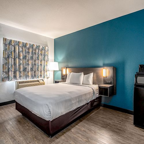 A modern hotel room with a double bed, blue accent wall, wall-mounted lights, mini fridge, microwave, and a desk with a chair.