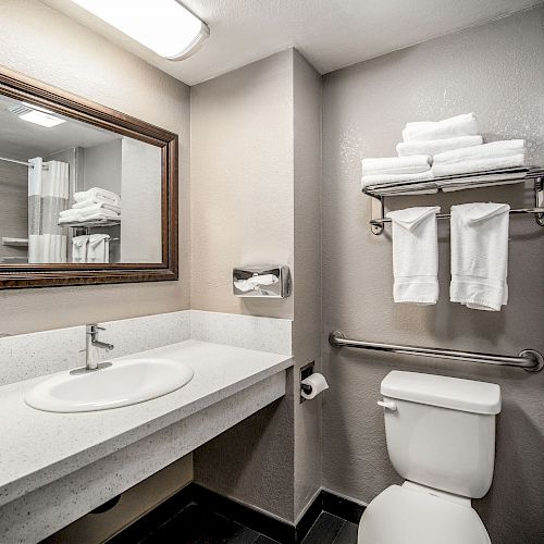 The image shows a clean, modern bathroom with a sink, mirror, toilet, and metal towel rack with folded white towels.