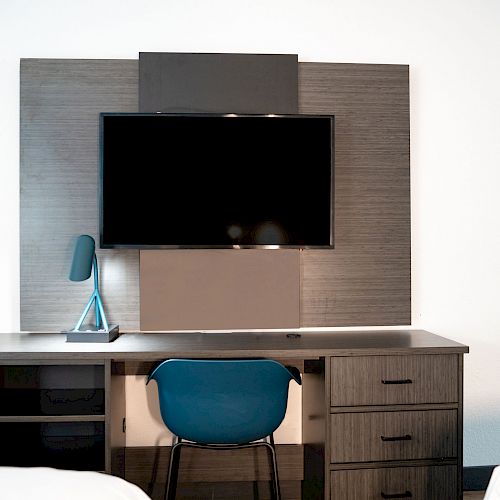 A desk set against a wall with a mounted TV, a blue chair, a desk lamp, and drawers is shown in the image.