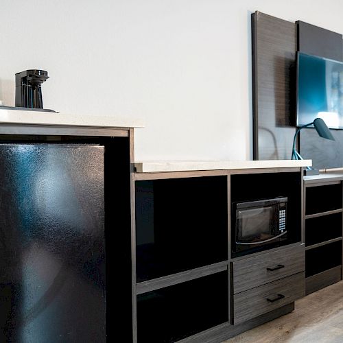 The image shows a modern workspace with a mini-fridge, coffee maker, desk, chair, mounted TV, and microwave against a plain white wall.