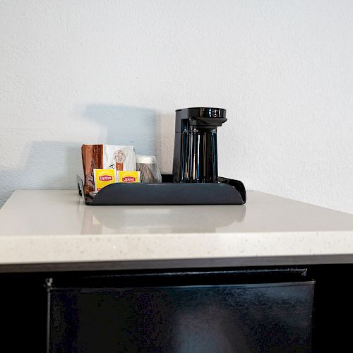 A small black coffee maker with a tray containing condiments, sugars, and stir sticks is placed on a white countertop.