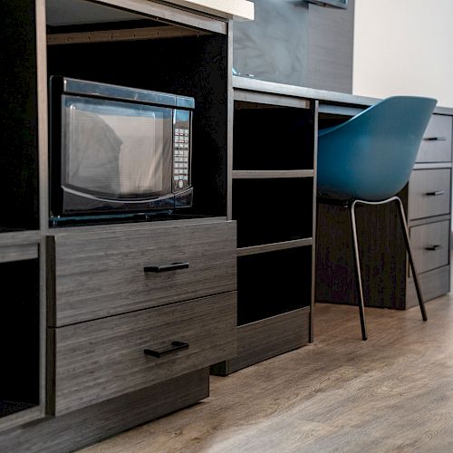 The image shows a desk area with drawers, a microwave on a shelf, and a modern blue chair, all on a hardwood floor.