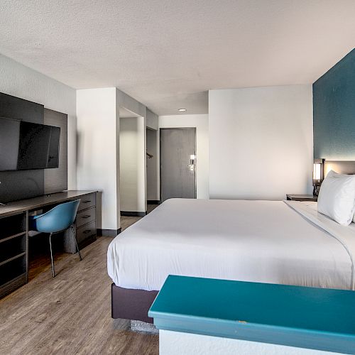 A modern hotel room with a large bed, TV, desk, chair, and bedside lamps, decorated in a minimalist style with blue and white accents.