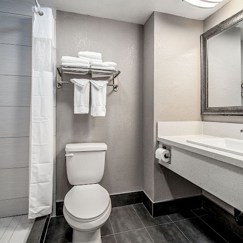 A clean bathroom with a toilet, shower, sink, a large mirror, and neatly folded towels on a shelf above the toilet, ending the sentence.