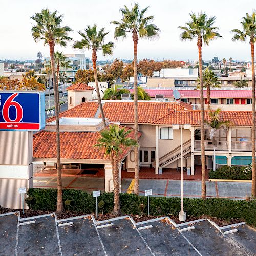 The image shows a Motel 6 with a red-tiled roof, surrounded by palm trees and adjacent parking, in an urban area.