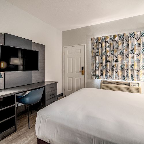 A hotel room with a bed, desk, chair, lamp, TV on the wall, and window with patterned curtains.