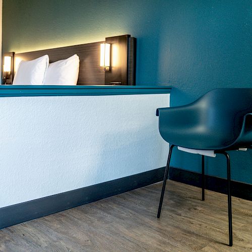 A modern hotel room features part of a bed with pillows, bedside lighting, a teal accent wall, and a teal chair on a wooden floor.