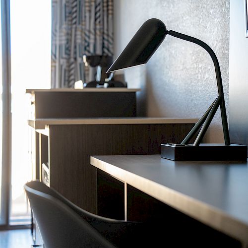 A modern desk with a black lamp, chair, and a window showing a balcony, in a room with minimalistic decor.