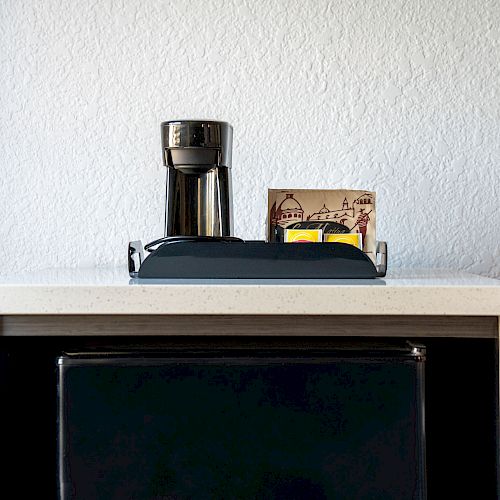 A coffee maker, a tray with a mug, and a box of coffee pods are on a counter above a mini refrigerator against a white wall.