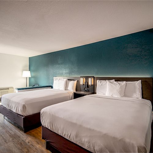 A hotel room with two neatly made beds, a teal accent wall, bedside lamps, a window, and a small air conditioning unit.