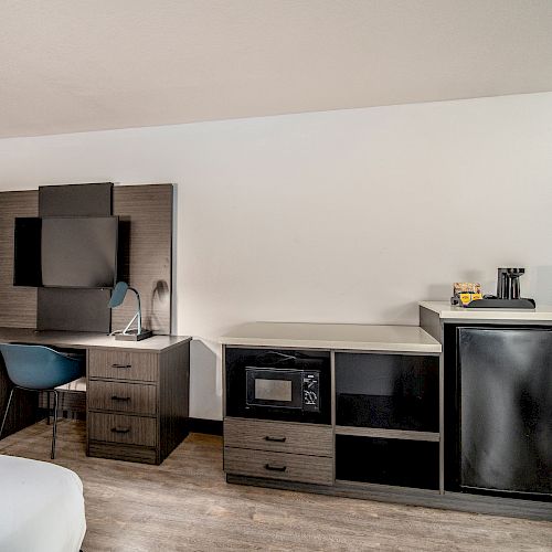 This image shows a room with a desk, office chair, wall-mounted TV, lamp, microwave, mini-fridge, and a small counter with a coffee maker.