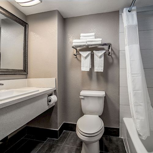 The image shows a bathroom with a modern design, featuring a sink, toilet, and shower-tub combo with a curtain. Towels are neatly stacked above.