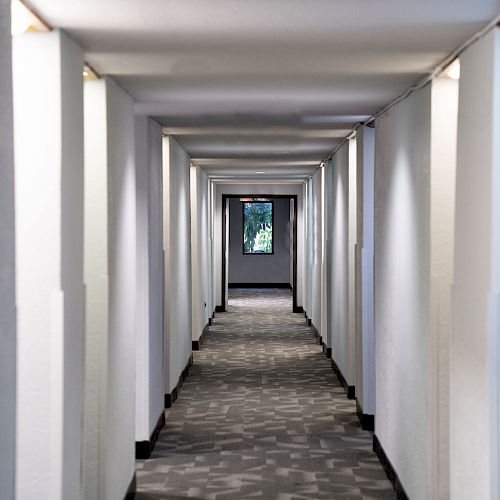 A long, narrow hallway with white walls and a patterned carpet leading to a window at the far end, allowing natural light to enter.