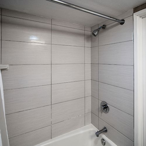 The image shows a clean bathroom with a white-tiled bathtub, a showerhead, a white curtain, and towels hanging to the left on a metal rack.