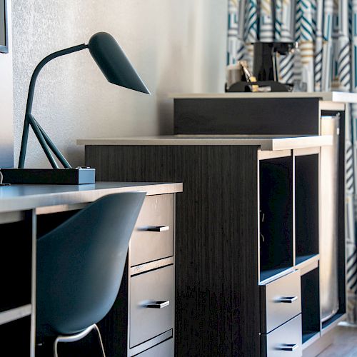 This image shows a modern workspace with a desk, chair, table lamp, and shelving units in front of geometric-patterned curtains, ending the sentence.