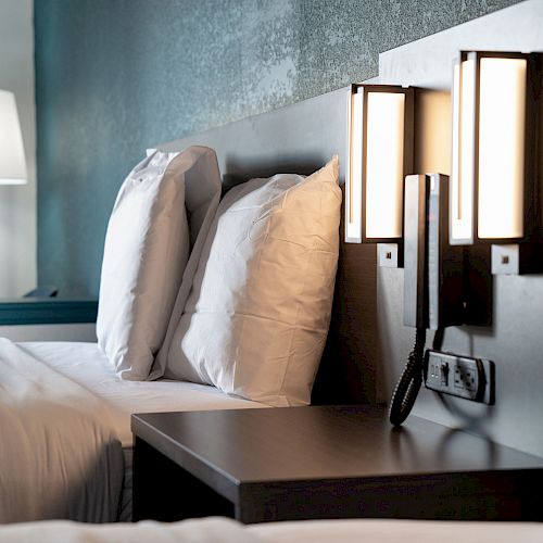 The image shows a neatly made bed with white pillows and linens, bedside table, modern wall-mounted lamps, and a floor lamp in a stylish bedroom.