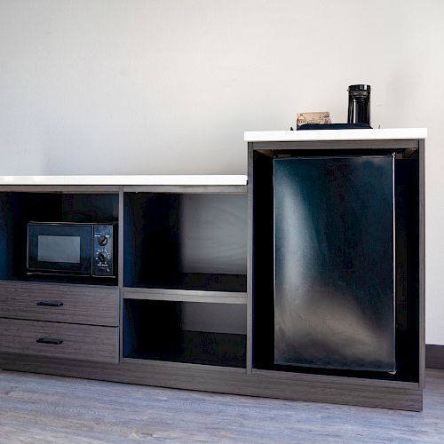 The image shows a modern kitchenette with a mini fridge, microwave, open shelves, and drawers. A coffee maker is on the countertop.