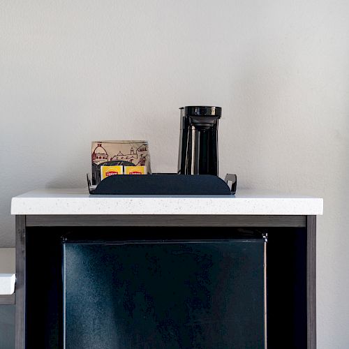 A small refrigerator with a white countertop. On top, there's a black coffee maker and a tray holding coffee supplies against a plain wall.