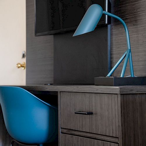 The image shows a modern desk setup with a blue chair and a matching blue desk lamp on a wooden desk next to a drawer and shelves.