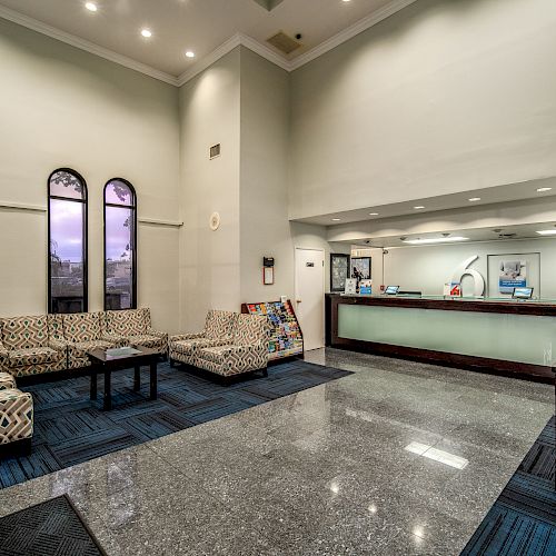 The image shows a spacious, modern lobby with a reception desk, seating area with patterned chairs, and a polished floor, under soft lighting.