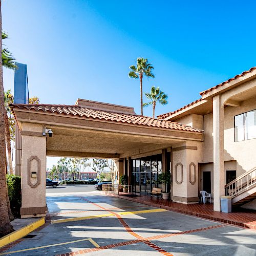 The image shows a building with a Spanish-style roof, an overhang for car drop-offs, palm trees, and a staircase. It has a sunny, clear day backdrop.