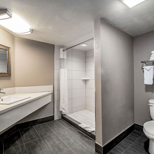 The image shows a clean, modern bathroom with a large mirror, sink, shower, toilet, and towel rack holding folded towels.