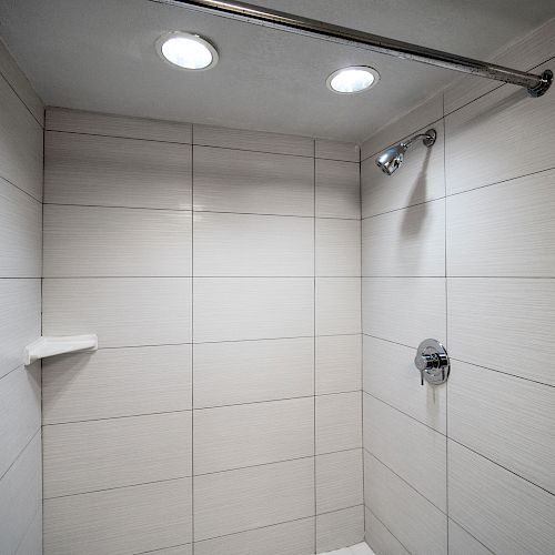 A modern tiled shower with two overhead lights, a showerhead, a soap shelf, and a partially visible shower curtain on the left side ending the sentence.