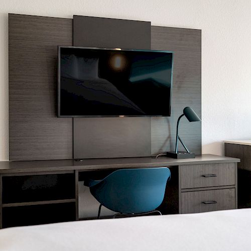 A modern hotel room setup featuring a wall-mounted TV, desk with a blue chair, lamp, and a small microwave or fridge on the right.