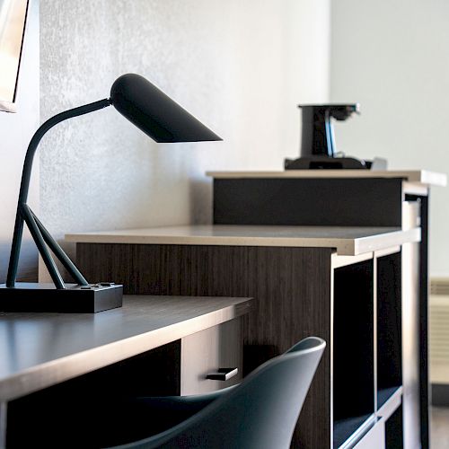 The image shows a modern desk setup with a sleek black lamp, office chair, and a coffee maker on another table in a minimalistic room.