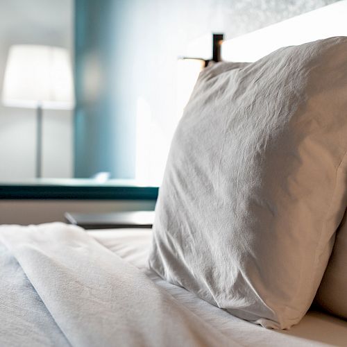 The image features a neatly made bed with white pillows and sheets, with a lamp and curtain in the background.