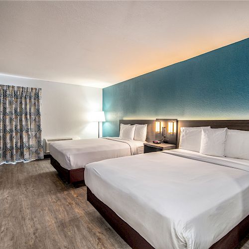 A hotel room featuring two queen-sized beds, a desk, a chair, a TV, and a lamp, with blue accent walls and curtains.