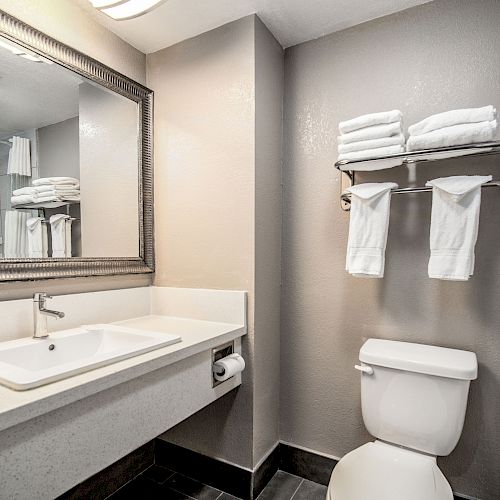 A clean bathroom with a large mirror, sink, toilet, a shelf with folded towels, and a shower curtain. The walls are light-colored.