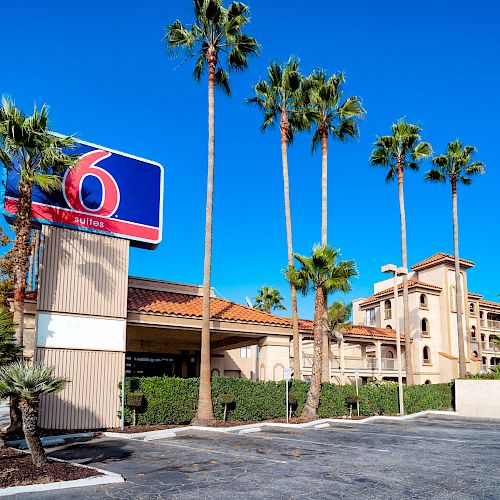 An exterior view of a Hotel 6 building with palm trees, a blue sky, and an empty parking lot.