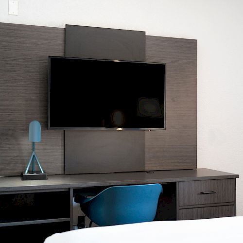 The image shows a modern desk setup with a black TV mounted on the wall, a blue table lamp, a blue chair, and drawers on the side, in a minimal room.