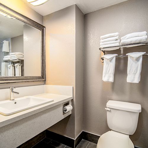 This image shows a clean, modern bathroom with a large mirror, sink, toilet, and neatly folded towels on a wall-mounted shelf above the toilet.