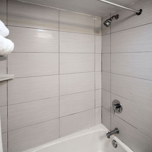 A bathroom featuring a white bathtub, a wall-mounted showerhead, a tiled wall, and a shelf with neatly folded white towels is shown in the image.