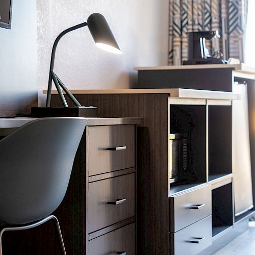 A modern, well-lit room with a desk, chair, lamp, shelving unit, coffee maker, and window with natural light coming through the blinds, creating a cozy atmosphere.